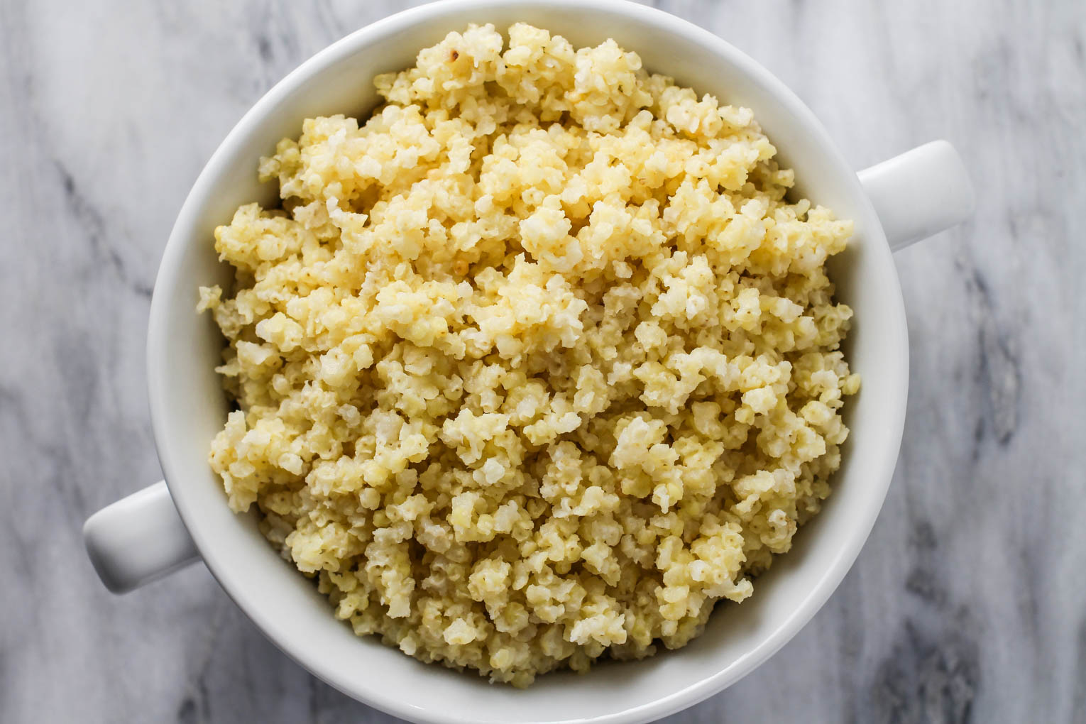 Overhead shot of cooked millet in a white bowl standing on a marble background.