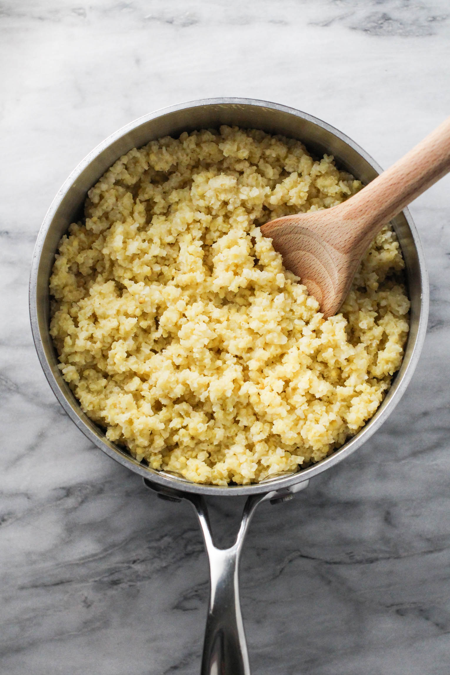 Cooked millet in a pot with a wooden spoon in it. The pot is standing on a marble background.