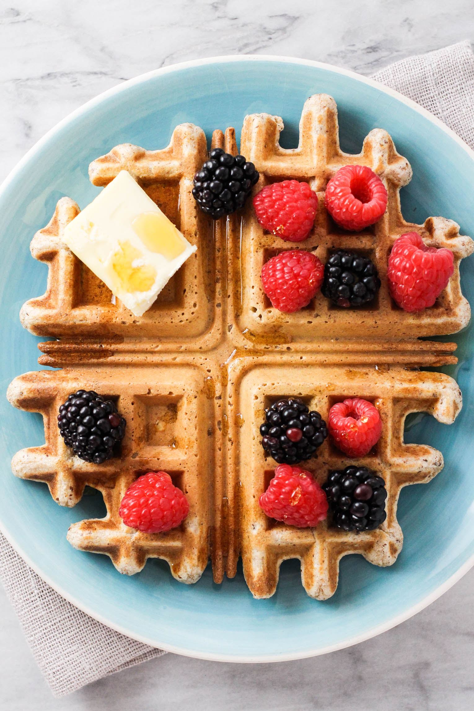 Overhead shot of a whole wheat waffle on a blue plate. The waffle is garnished with berries, butter, and maple syrup. 