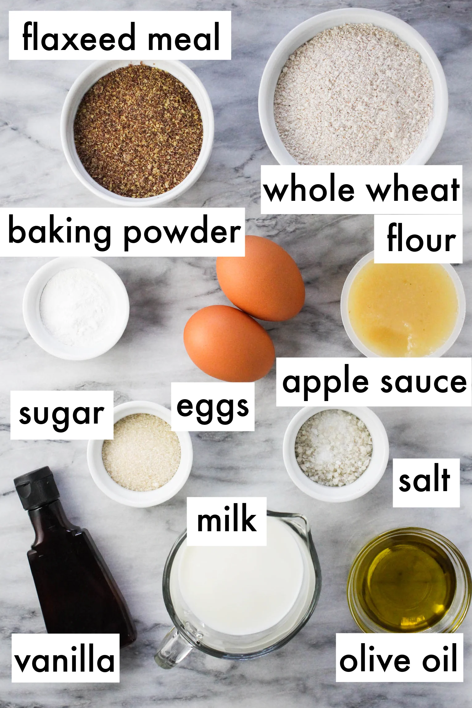The ingredients for the recipe dispayed on marble background. The ingredients are labeled as follows: flaxeed meal, whole wheat flour, baking powder, sugar, eggs, apple sauce, vanilla, milk, olive oil.