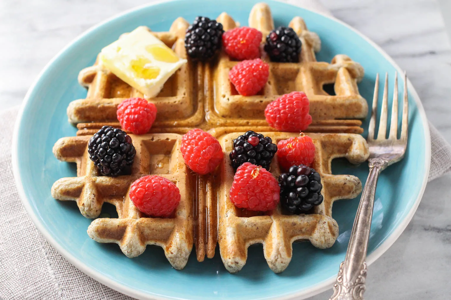 Whole wheat waffle on a blue plate, The waffles is garnished with berries, butter, and maple syrup. A silver fork is to the right of the waffle.