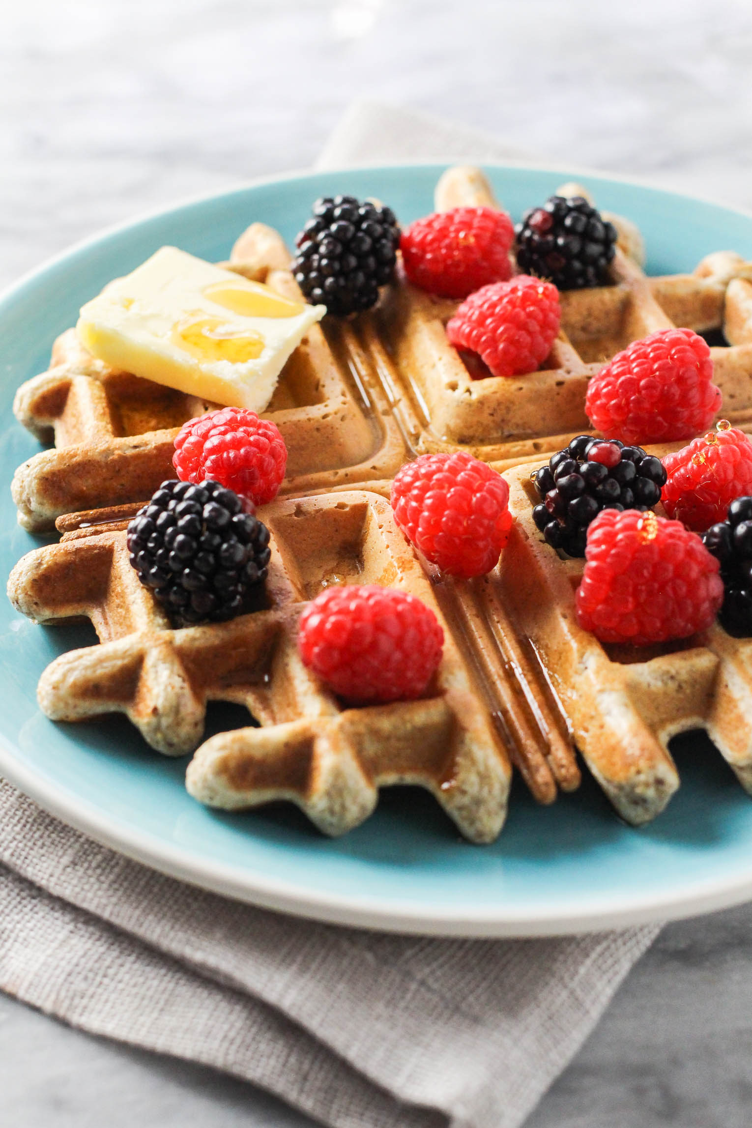 A side view shot of a whole wheat waffle on a blue plate. The waffle is garnished with berries, butter, and maple syrup.