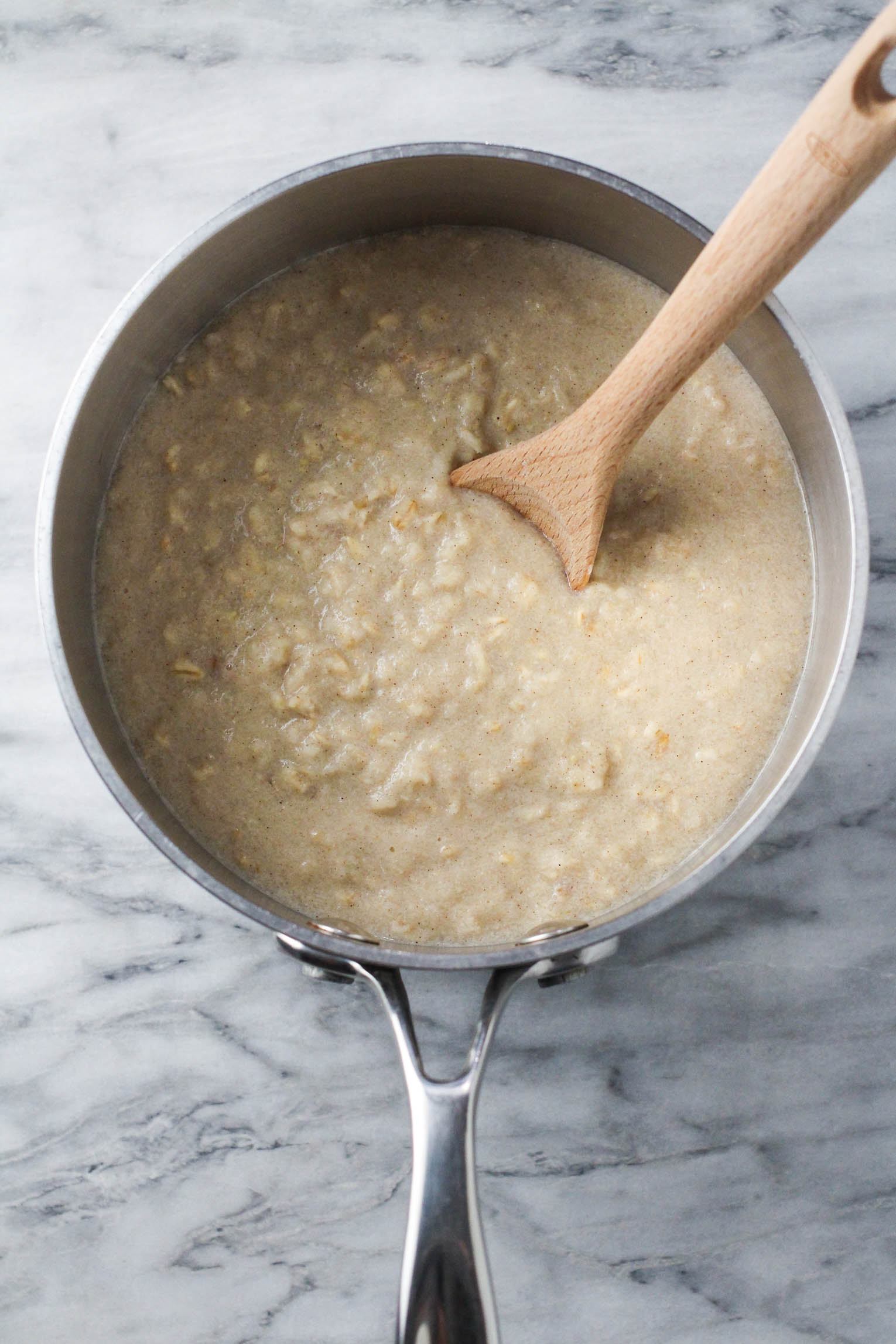 Overhead shot of oatmeal in a saucepan standing on a marble background. A wooden spoon is in the oatmeal.