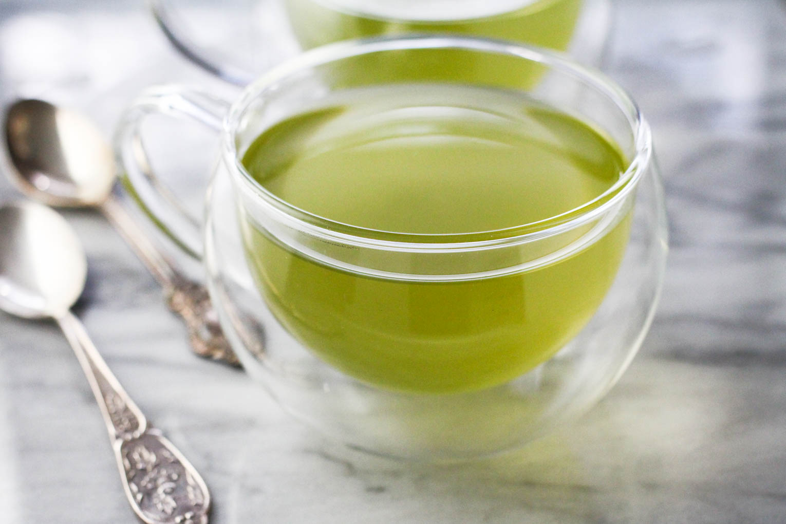 Close up side view shot of green tea and a glass tea cup with silver spoons to the left.