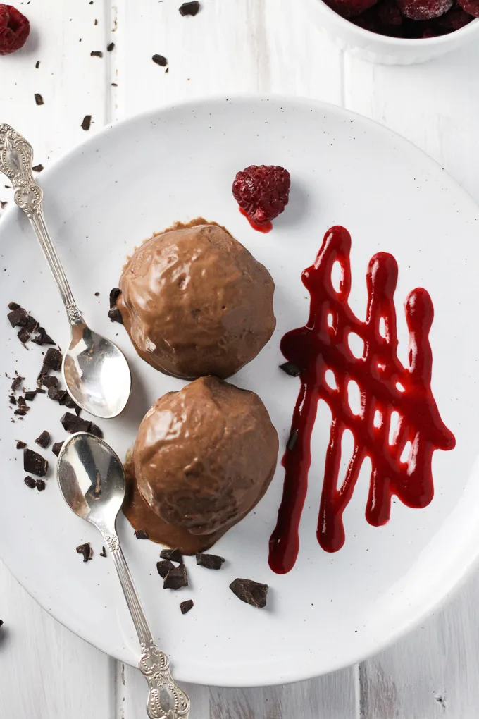 Two scoops of chocolate ice cream on a plate. Two silver spoons to the left. Drizzle of raspberry sauce to the right.