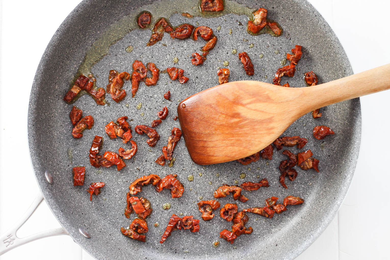 Overhead shot of sun-dried tomatoes in olive oil in a skillet with a wooden spatula.