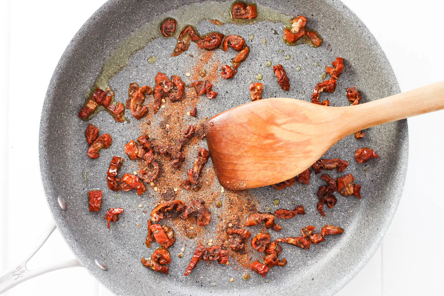Overhead shot of sun-dried tomatoes being mixed with spices with a wooden spatula.