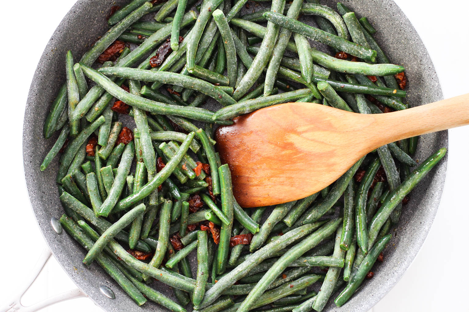 Frozen green beans in a skillet being mixed with sun-dried tomatoes with a wooden spatula.