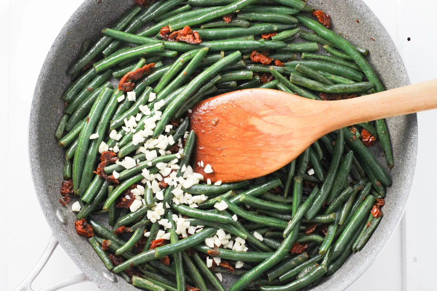 Green beans being tossed with garlic with a wooden spatula.