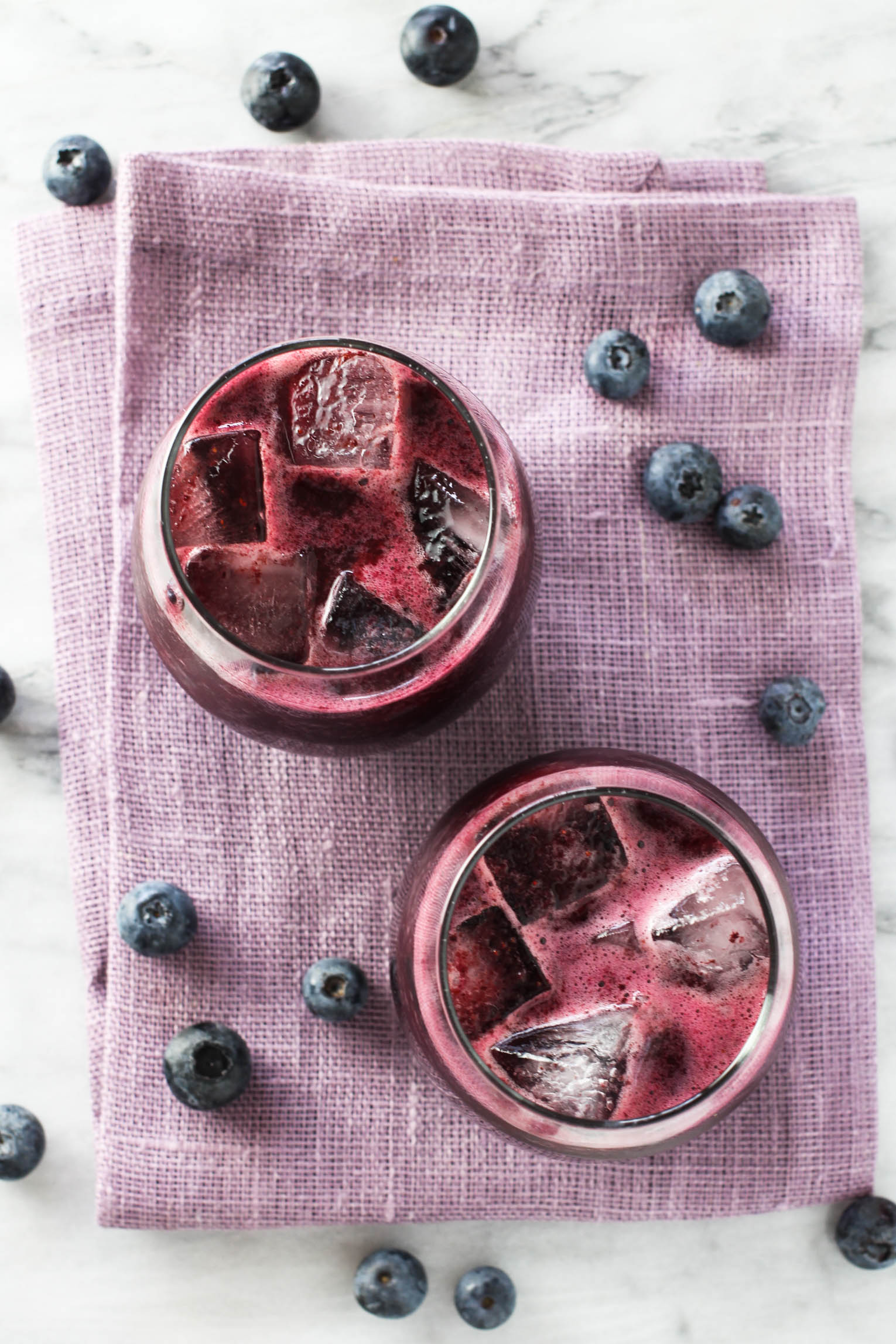Overhead shot of two glasses with blueberrie juice with ice. The glasses are standing in a purple napkin. There are blueberries scattered around the glasses.