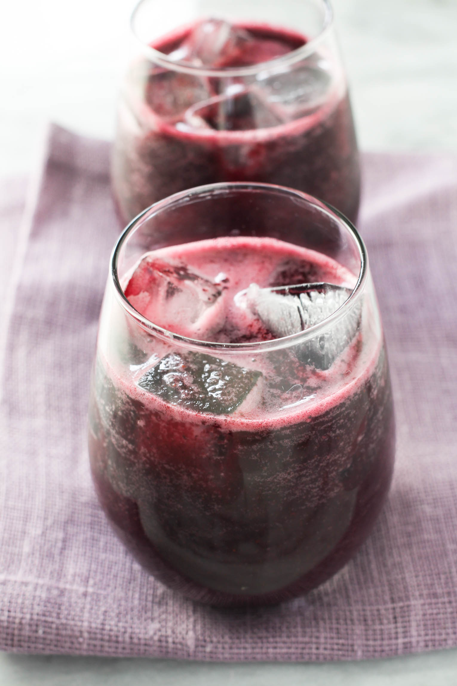 A glass with blueberry juice and ice standing on purple napkin. There is another glass with blueberry juice in the backgraound.