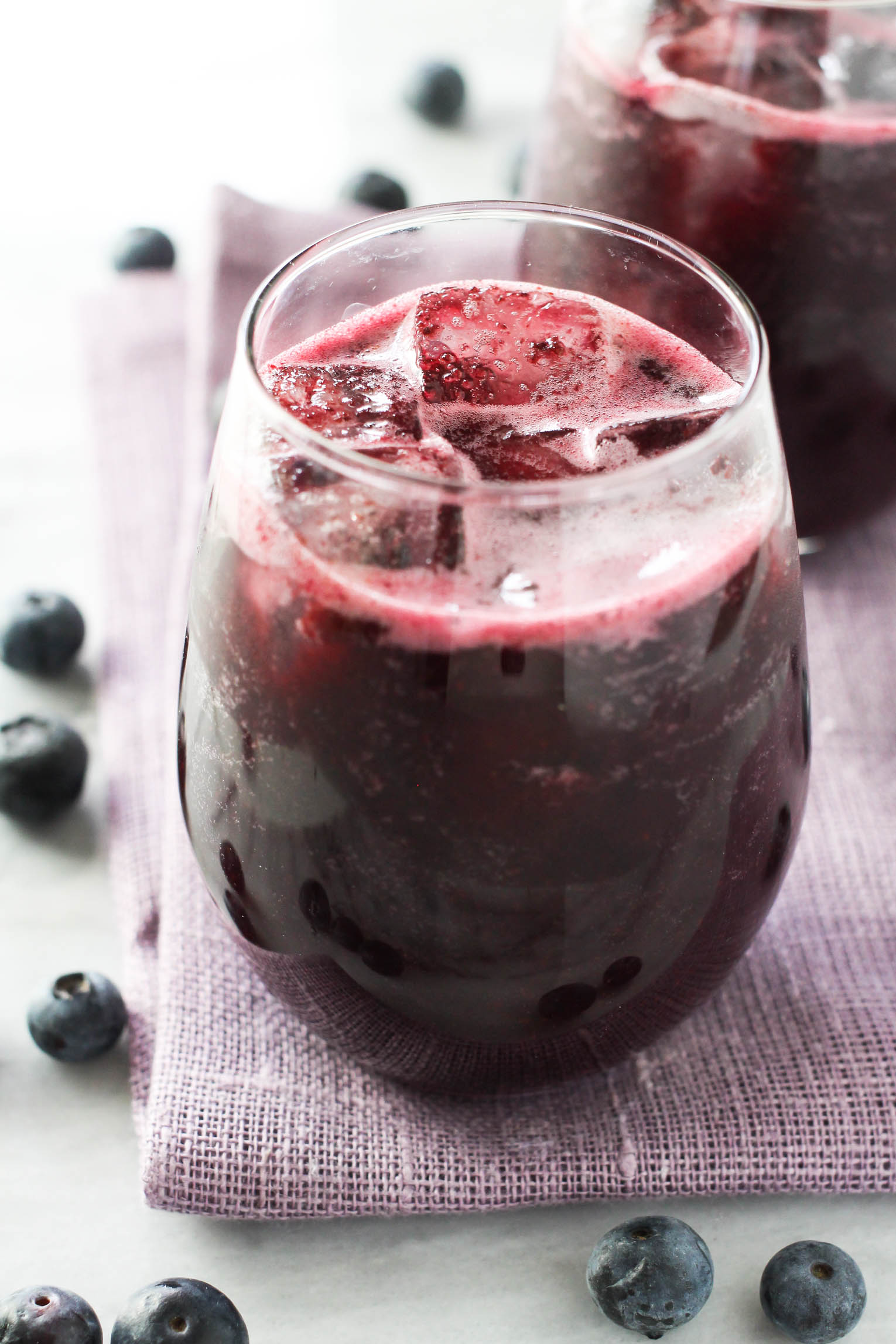 A glass with blueberry juice with ice cubes. The glass is standing on a purple napkin. There are fresh blueberries around the glass. 