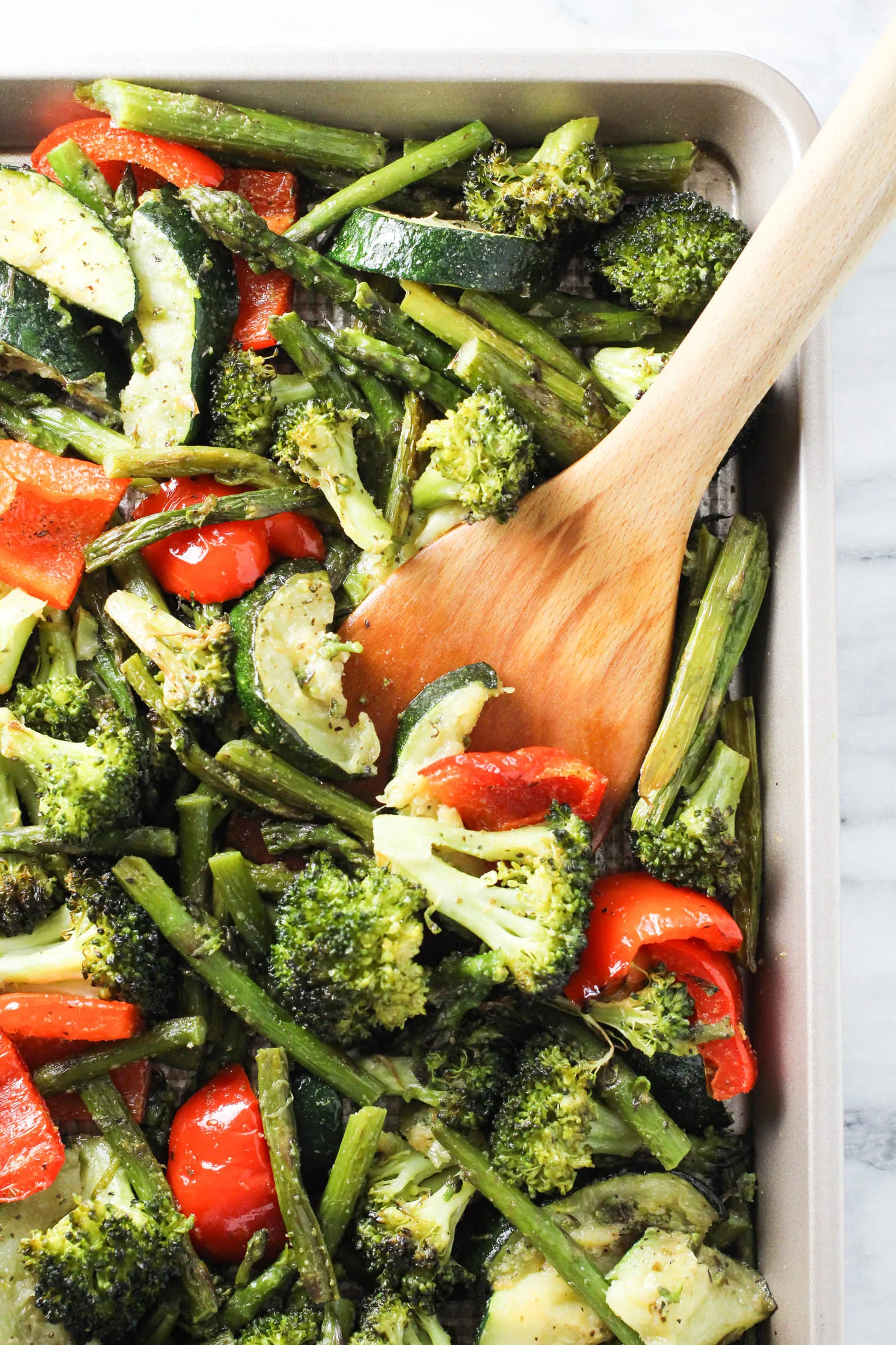 Roasted mixed vegetables on a baking sheet being scooped with a wooden spatula.
