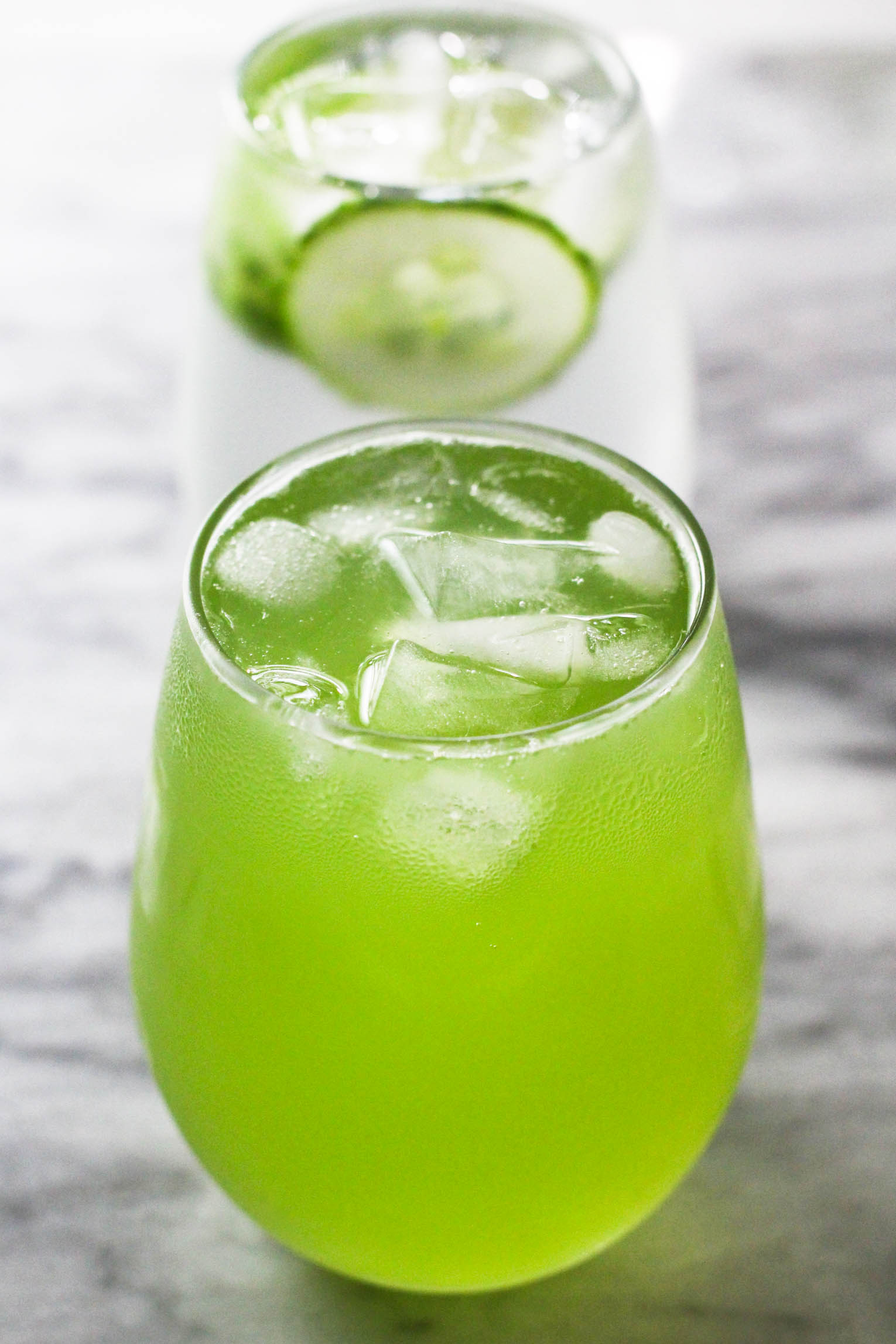 A glass with cucumber juice water and ice cubes. A glass with water with cucumber slices in the background.