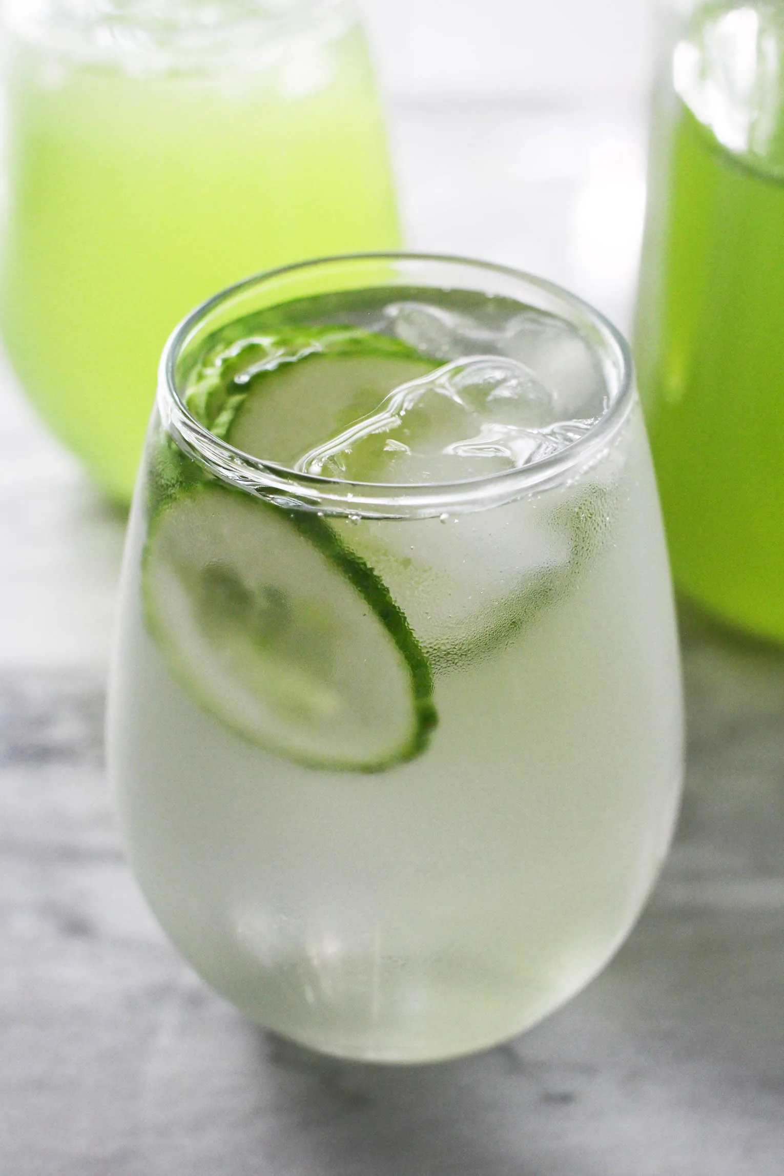 A glass with water, cucumber slices, and ice. Another glass with green liquid in the bakcground.