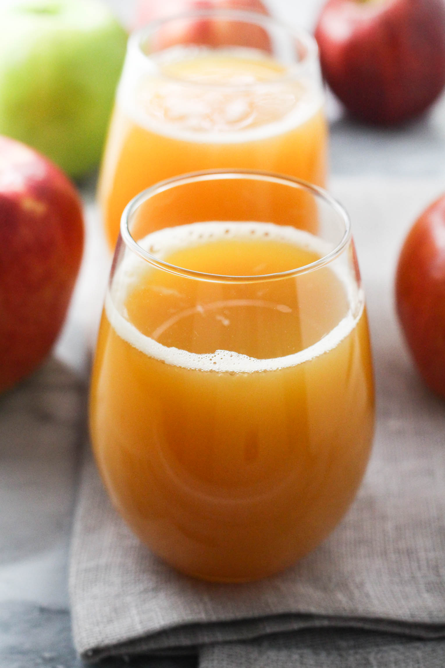 A glass with apple juice. There is another glass with juice behind it and some apples in the background.