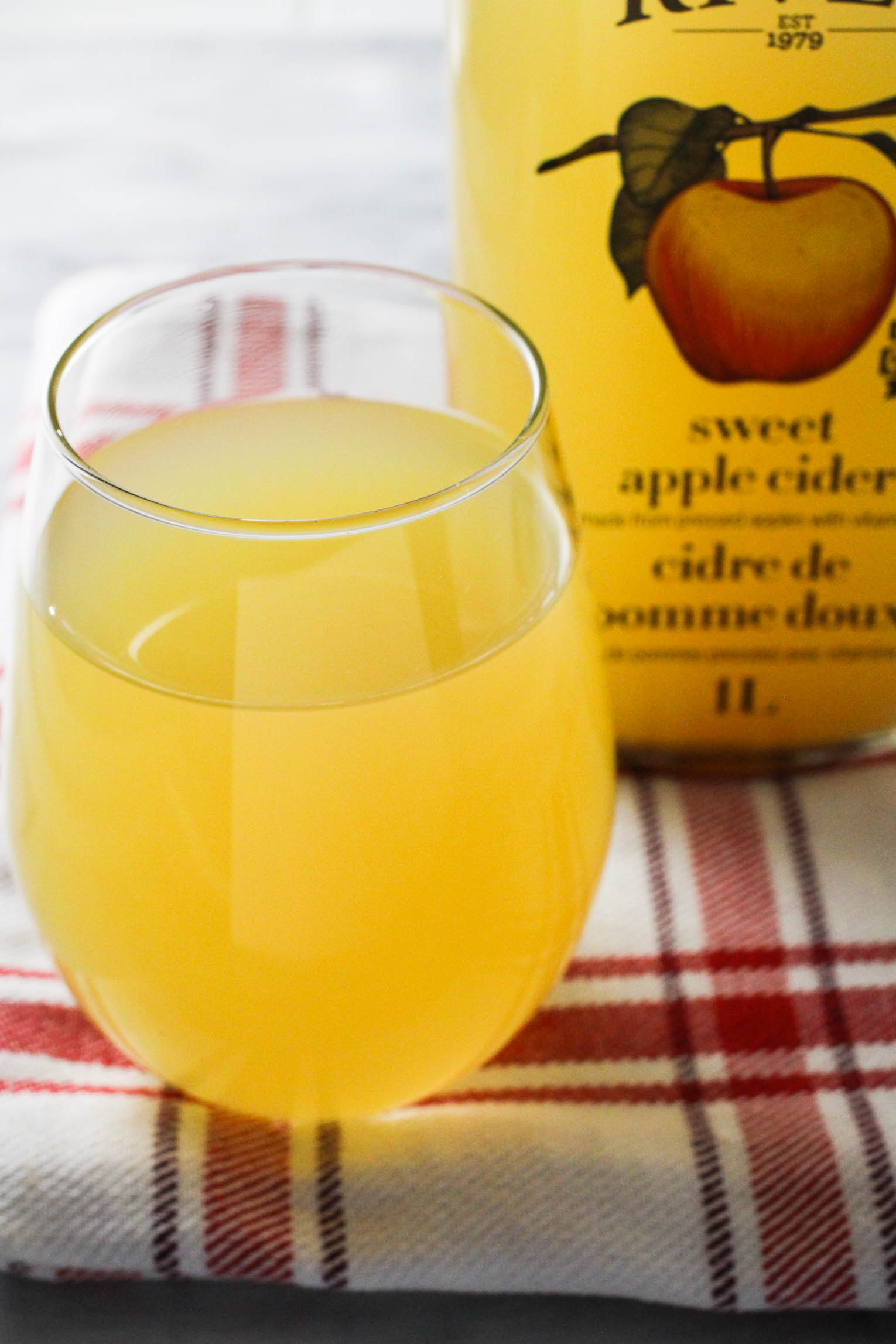 Apple cider in a glass standing on tea towel. A bottle with apple cider in the background.