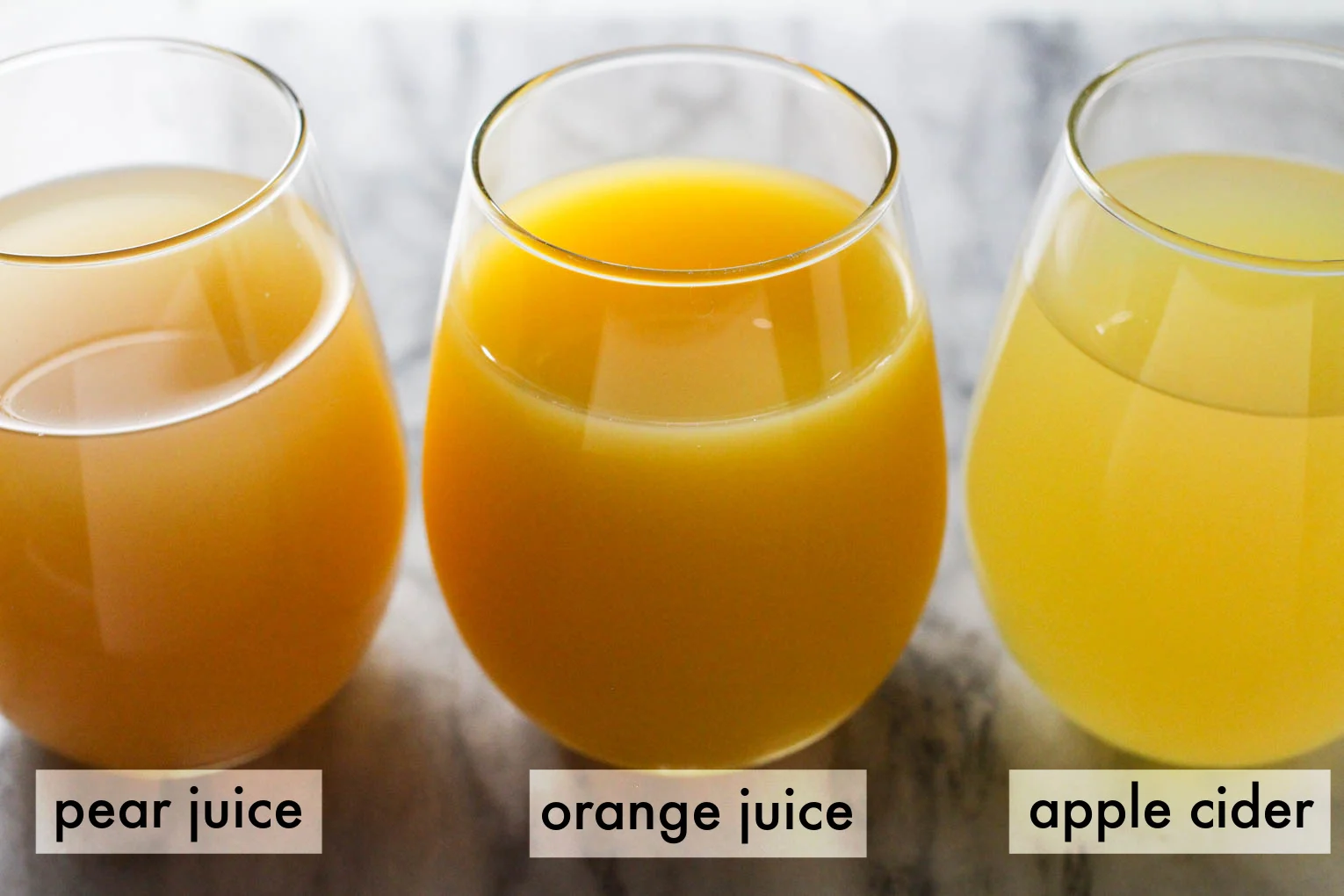 Three glasses with fruit juices standing on marble background. Below each glass, there is a text overlay as follows: pear juice, orange juice, apple cider.