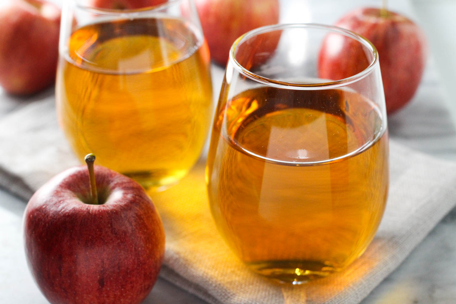 Two glasses with apple juice standing on a napkin. One apple in front of the glasses and a few behind the glasses.