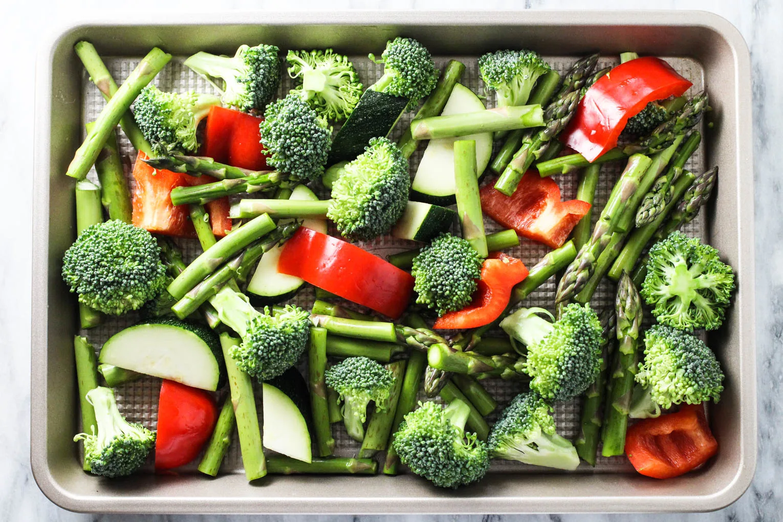Sliced broccoli, zucchini, red bell pepper, and asparagus spread on a baking sheet. 