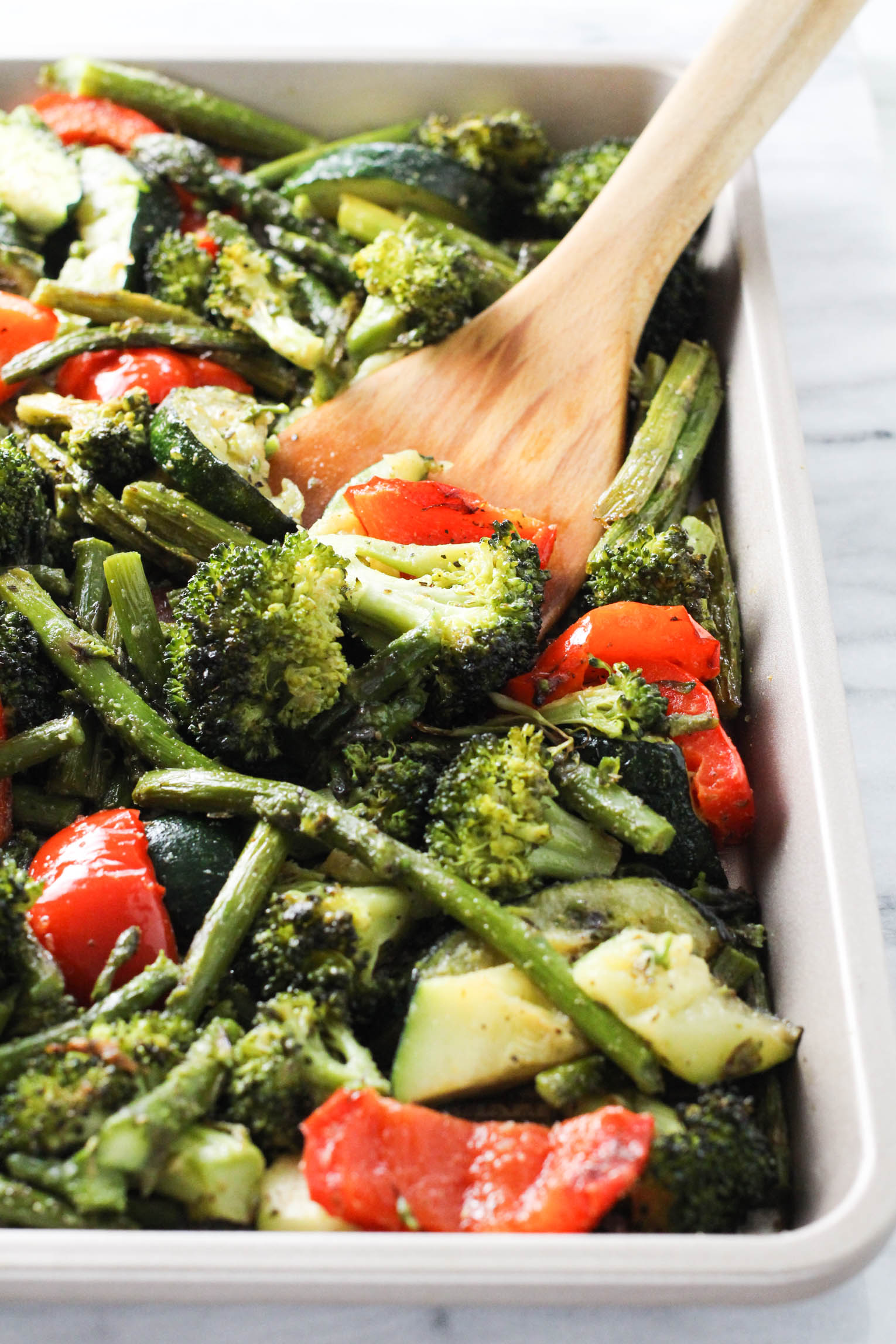 Roasted mixed vegetables on a baking sheet, being scooped with a wooden spoon.