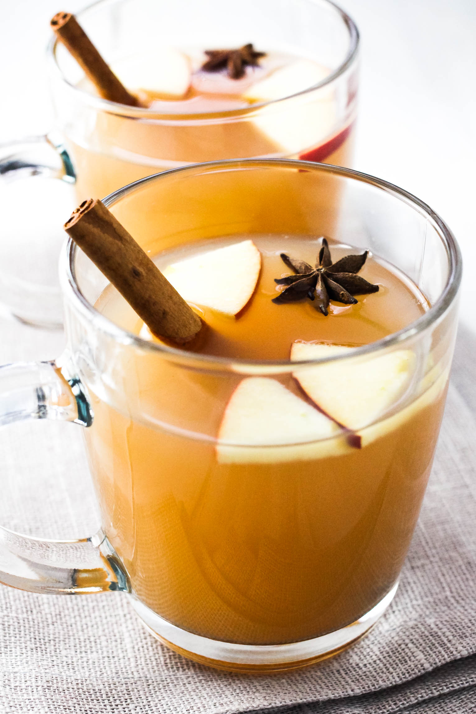 Two mugs with hot apple cider garnished with cinnamon sticks, apple slices and anise star.