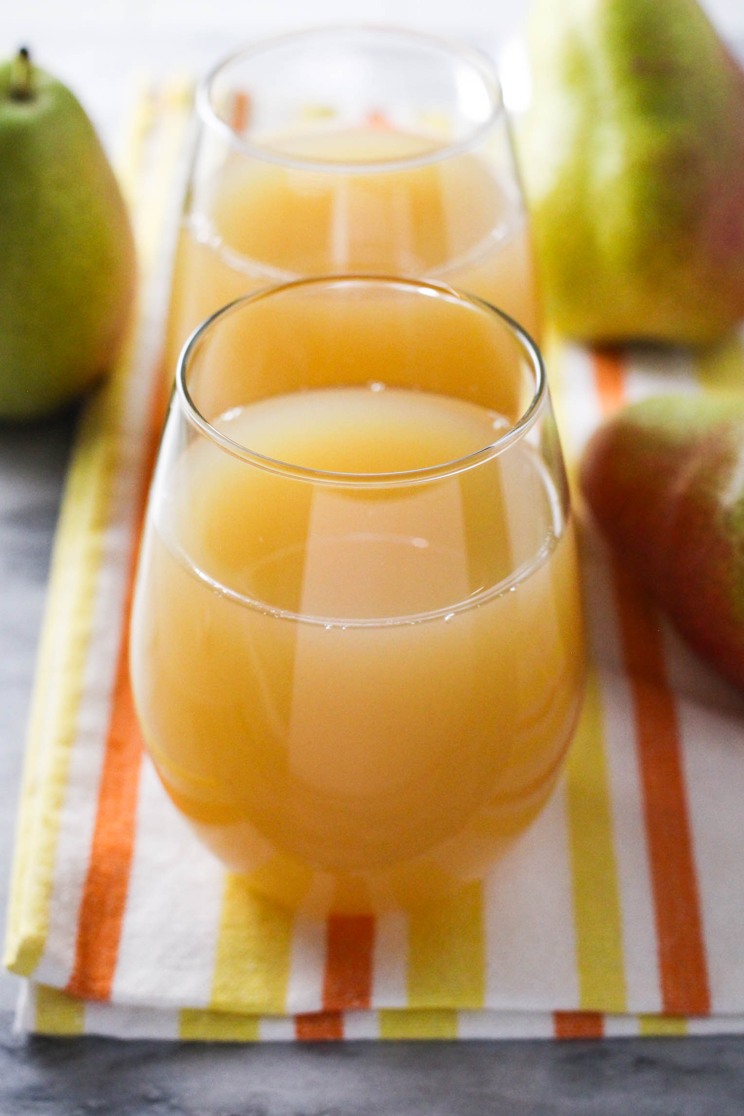 Two glasses with pear juice standing on a tea towel. Three pears beside the glasses.