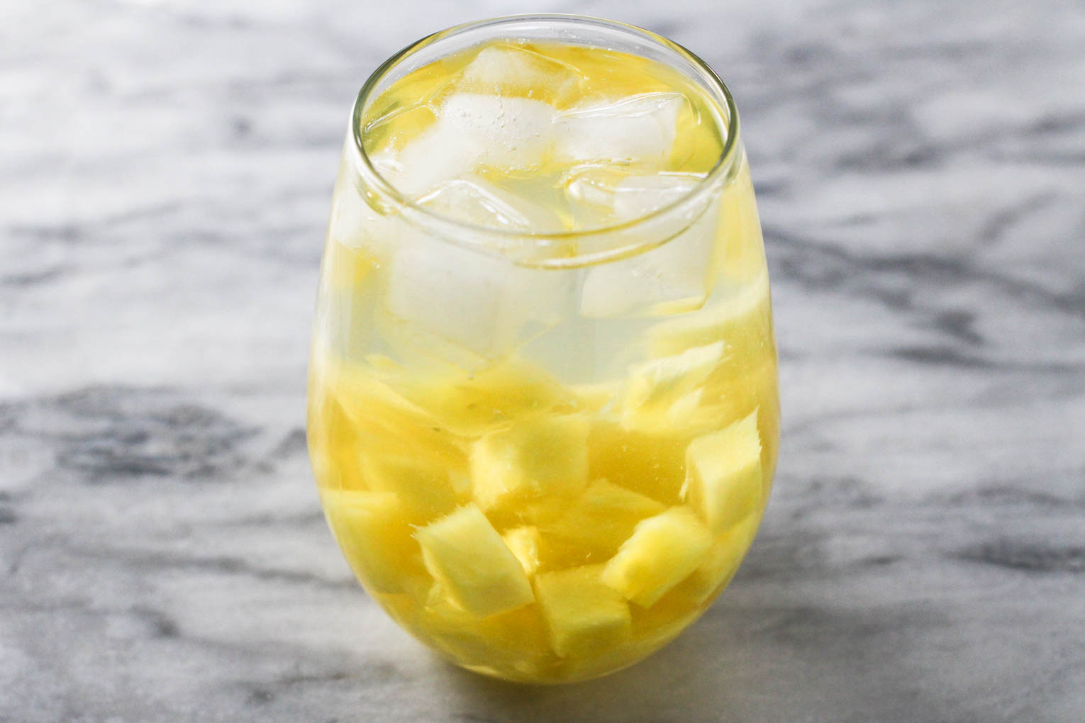 Close up shot of a glass of pineapple water with ice cubes and small pieces of pineapple. The glass is standing on a marble background. 