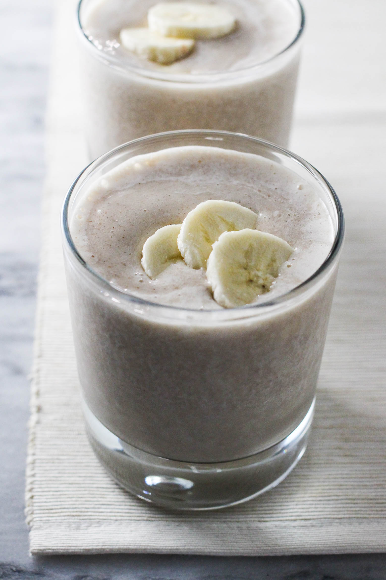 A glass of banana smoothie garnished with three banana slices. Another glass in the background.