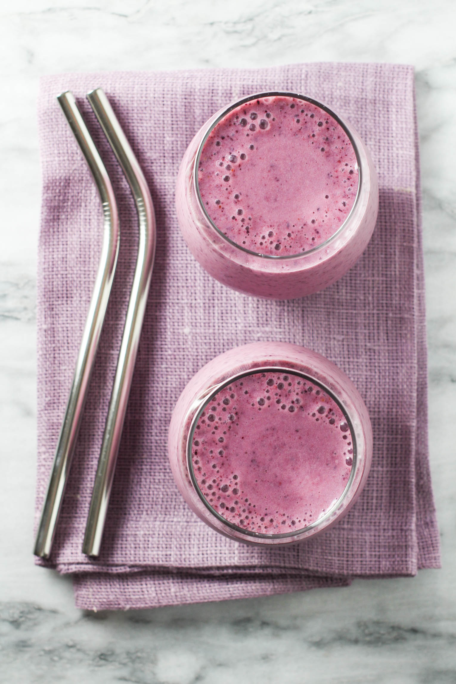 Overhead shot of two smoothie glasses standing on a tea towel. Two metal drinking straws to the left.