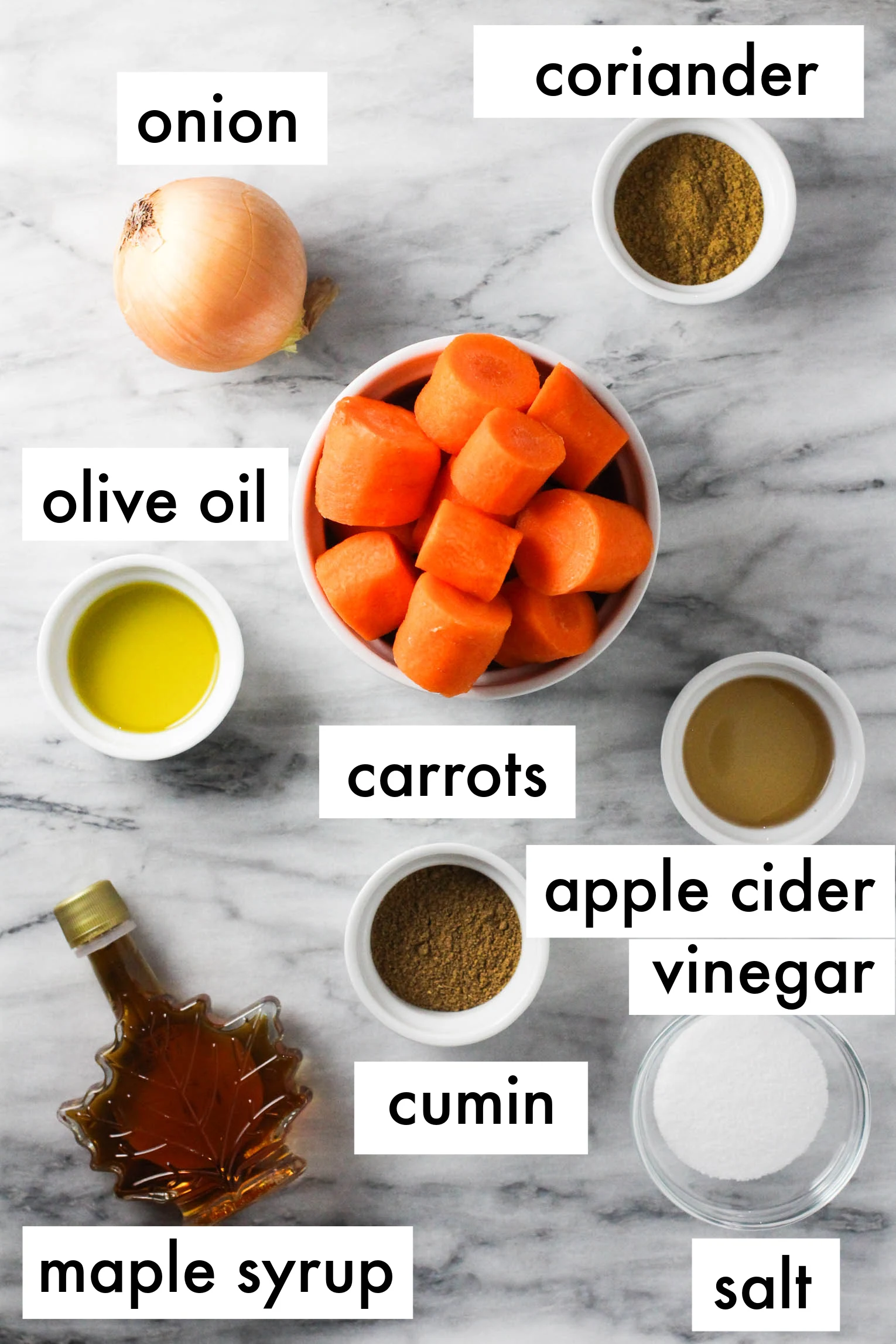 Carrot mash ingredients on marble background. The ingredients are labeled as follows: onion, coriander, olive oil, carrots, apple cider vinegar, cumin, maple syrup, salt.