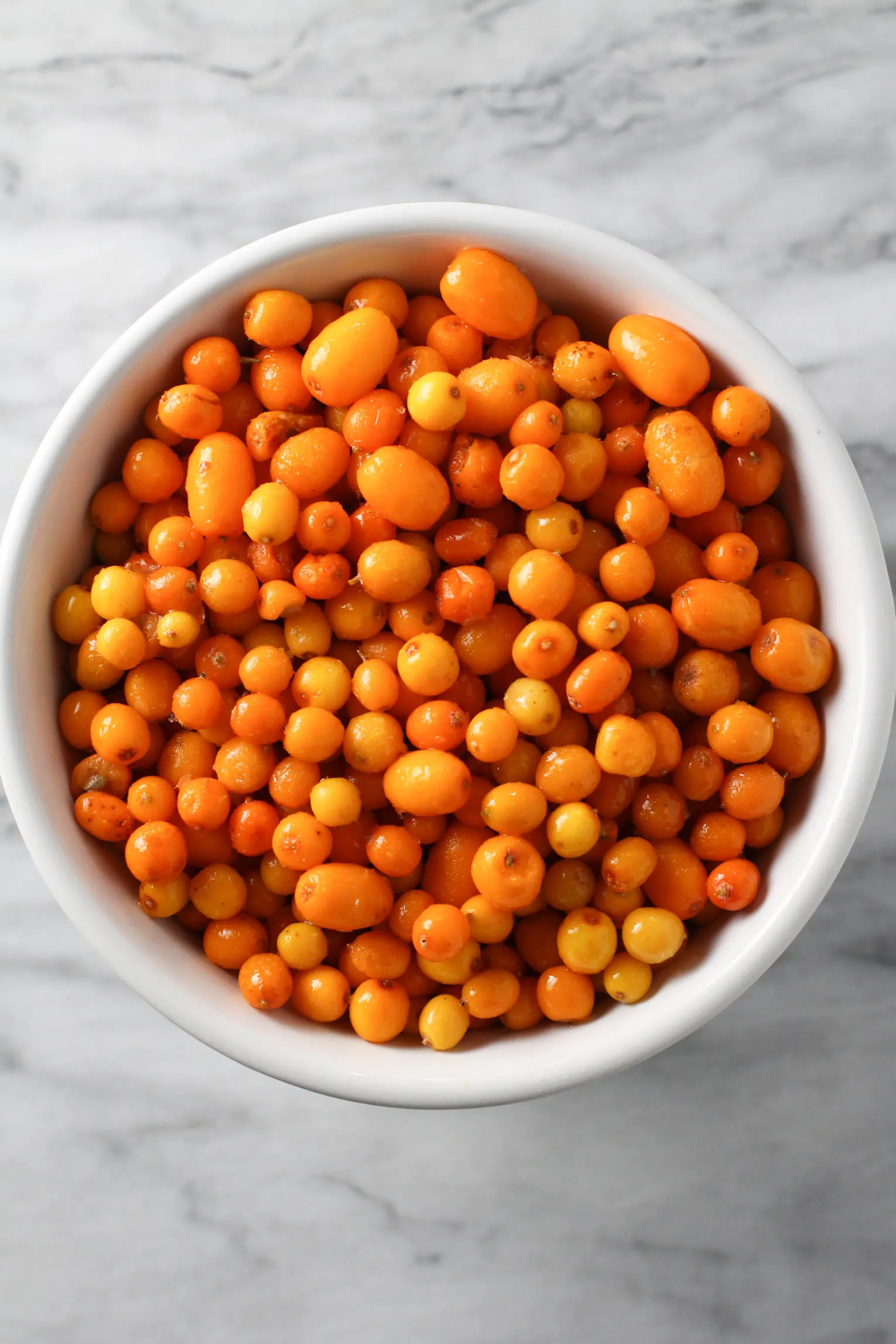 Overhead shot of sea buckthorn berries in a white bowl standing on a marble background.