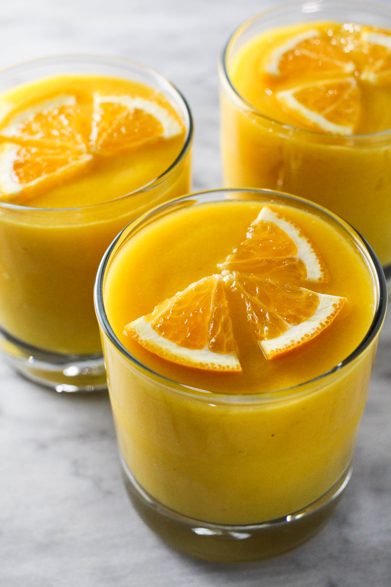 Three glasses of orange mango smoothie standing on marble background. The smoothies are garnished with orange slices.