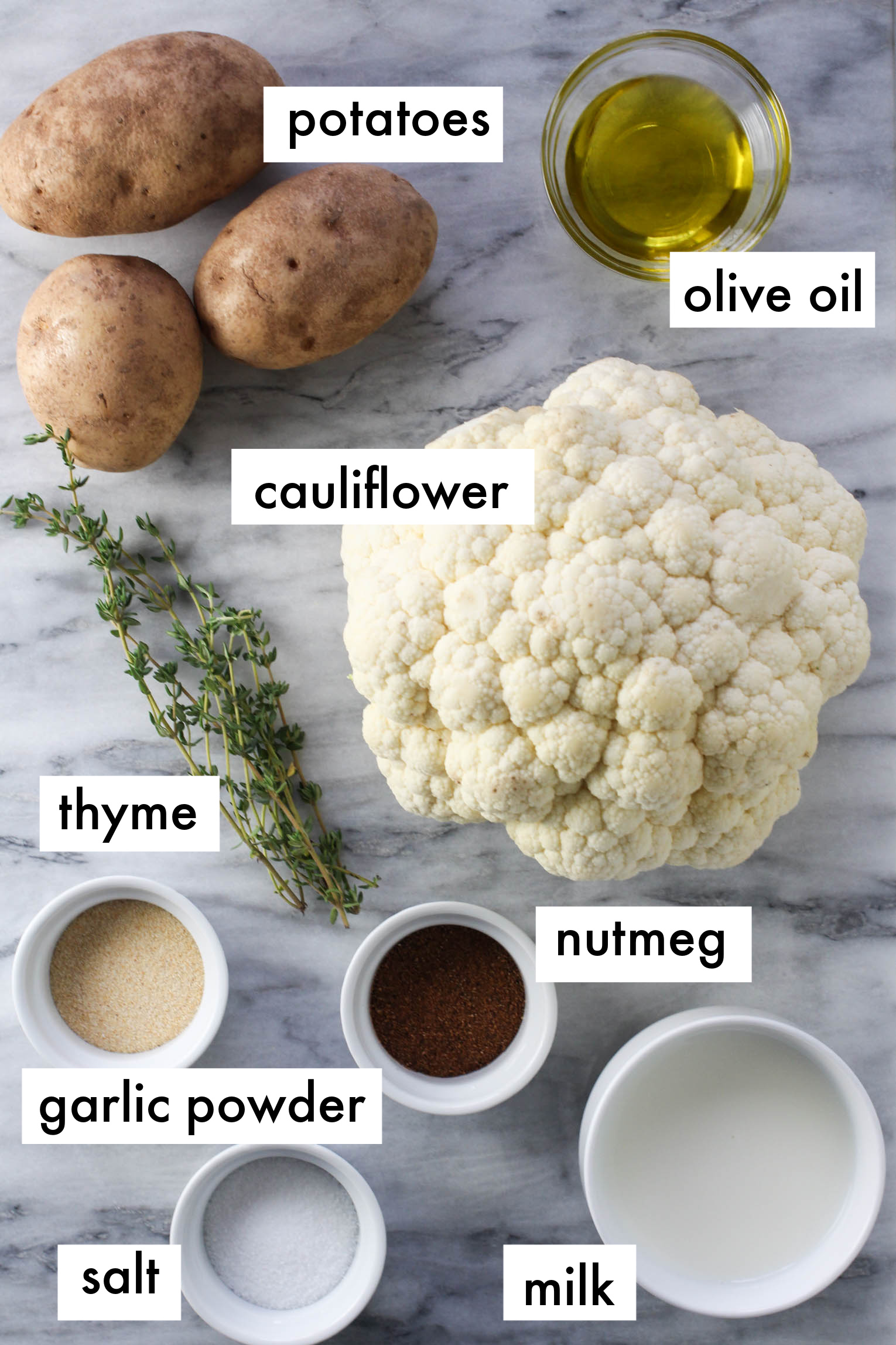 Ingredients for potato and cauliflower mash displayed on marble background. The ingredients are labeled as follows: potatoes, olive oil, cauliflower, thyme, nutmeg, garlic powder, salt, milk.