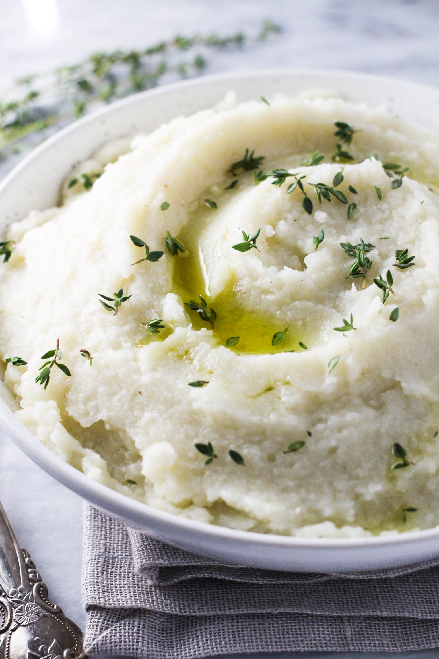Mashed potatoes and cauliflower in awhite bowl standing on a napkin. The mash is garnished with olive oil and fresh thyme leaves.