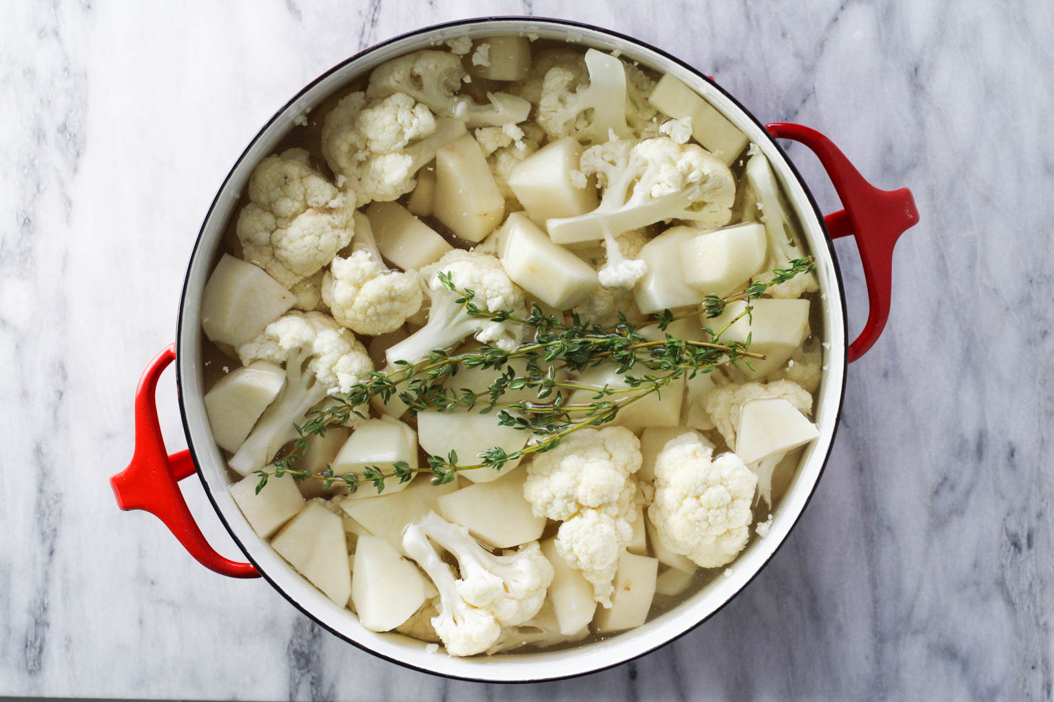 Potato chunks, cauliflower florets, and thyme sprig in a pot standing on marble background.
