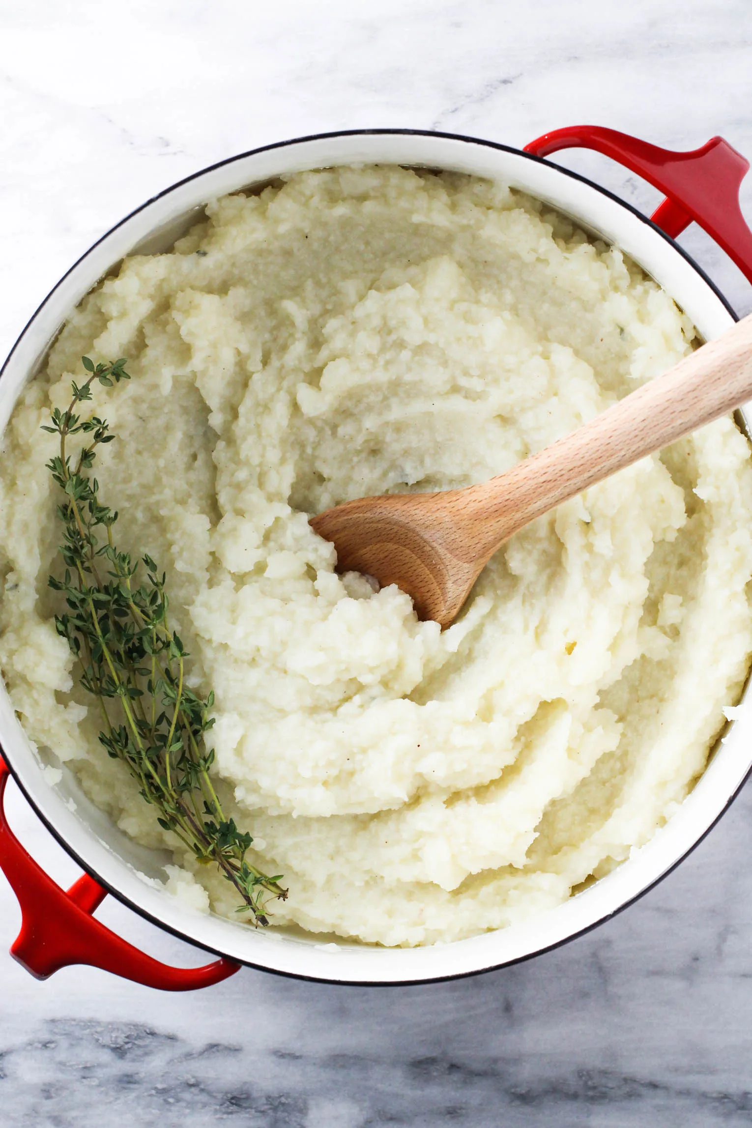 Potato and cauliflower mash in a pot with a wooden spoon in it. Thyme sprigs on the left side of the mash.