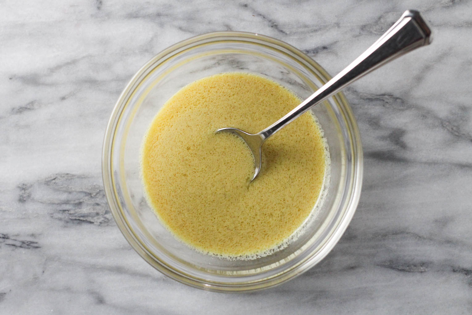 Dijon mustard vinaigrette in a small bowl standing on a marble background. There is a spoon in the bowl.