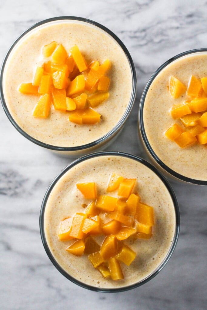 Overhead shot of three glasses of peach banana smoothie on marble background. The smoothies are garnished with diced peaches.