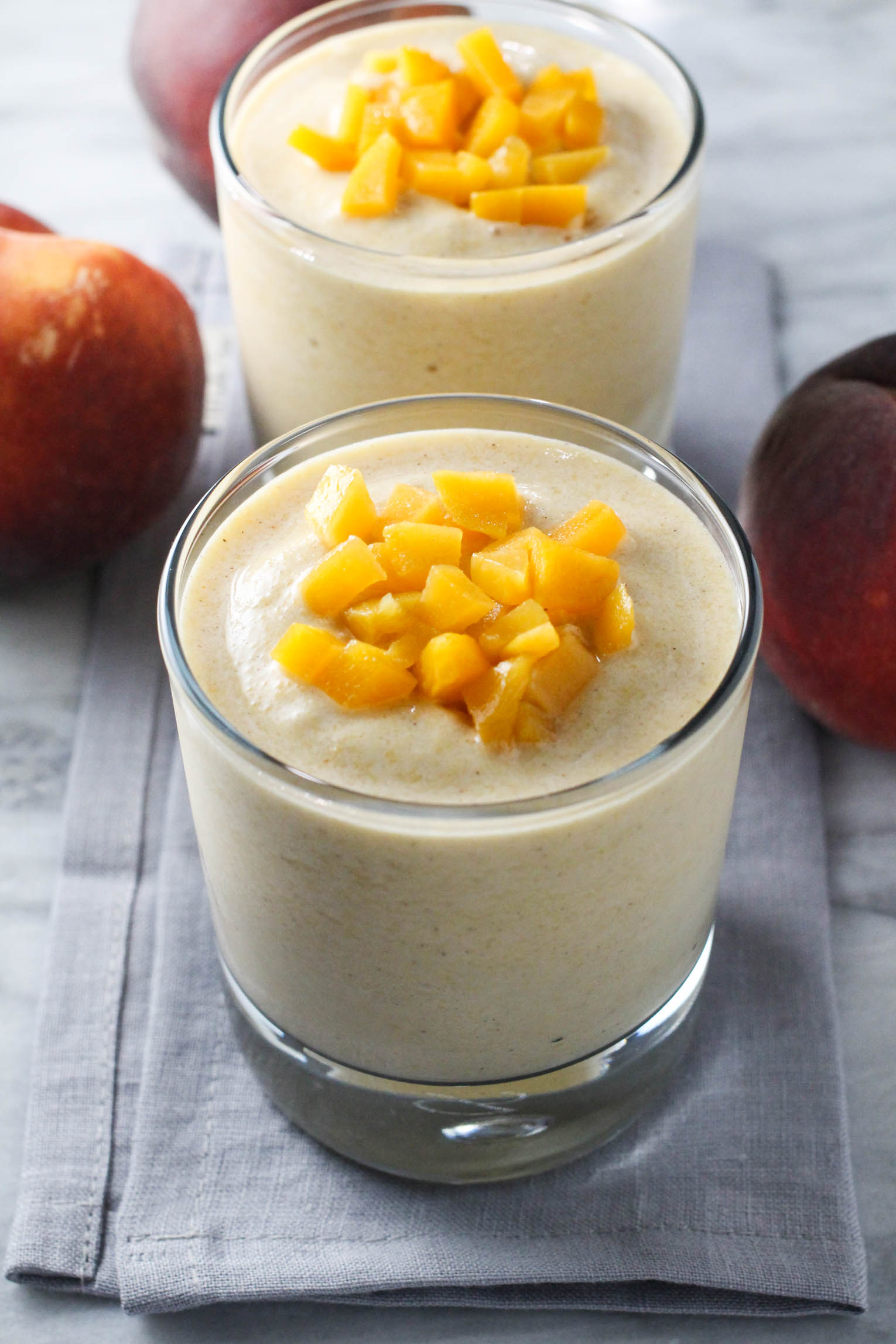 Two glasses of peach banana smoothie standing on a gray napkin. Three peaches in the background.