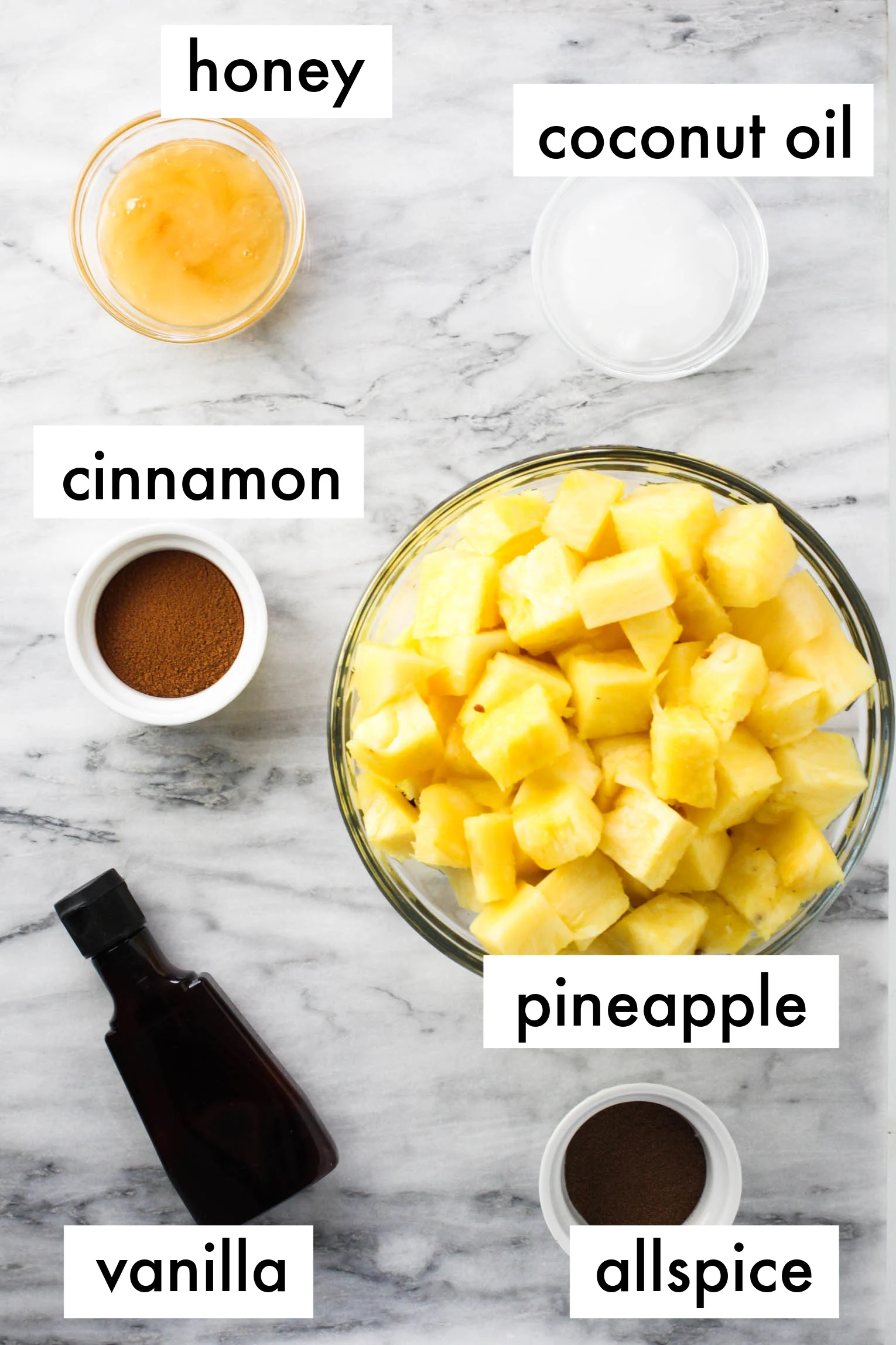 Roasted pineapple ingredients displayed on the marble background. The ingredients are labeled as follows: honey, coconut oil, cinnamon, pineapple, vanilla, allspice.