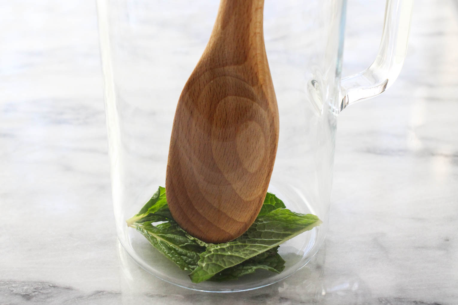 Mint leaves in a pitcher being muddled with a wooden spoon.