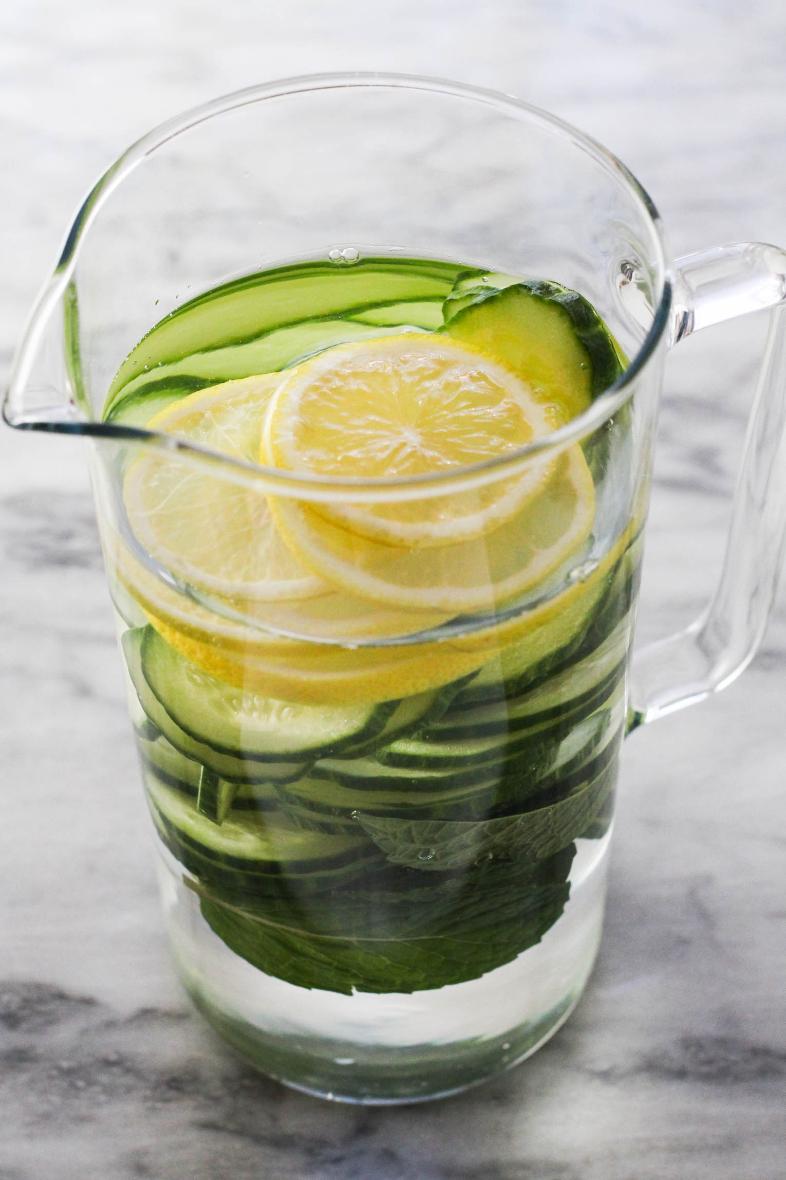 Mint leaves, cucumber slices, lemon slices, and water in a pitcher standing on marble background.