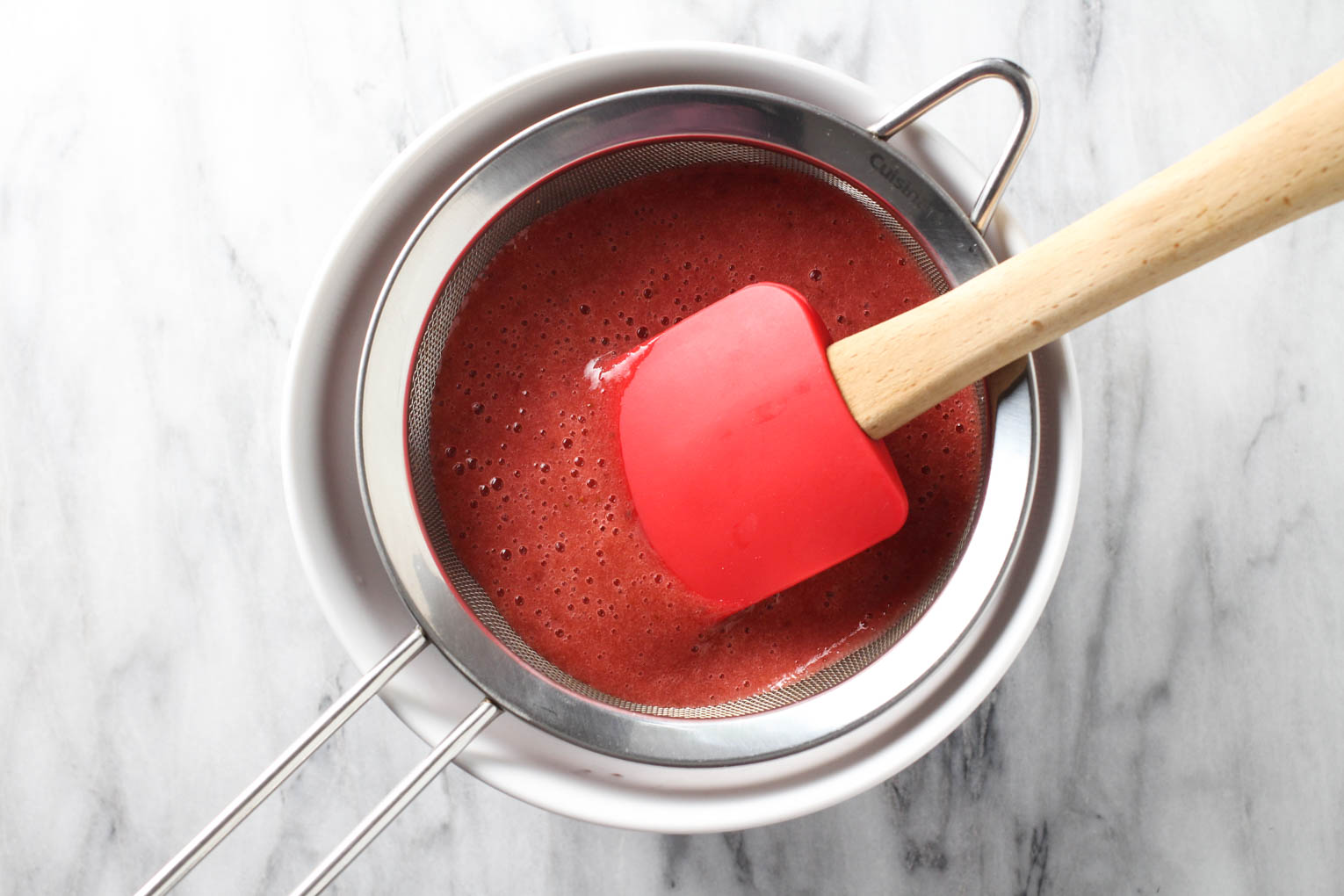 Overhead shot of strawberry sauce being pushed through a fine-mesh sieve with a rubber spatula.