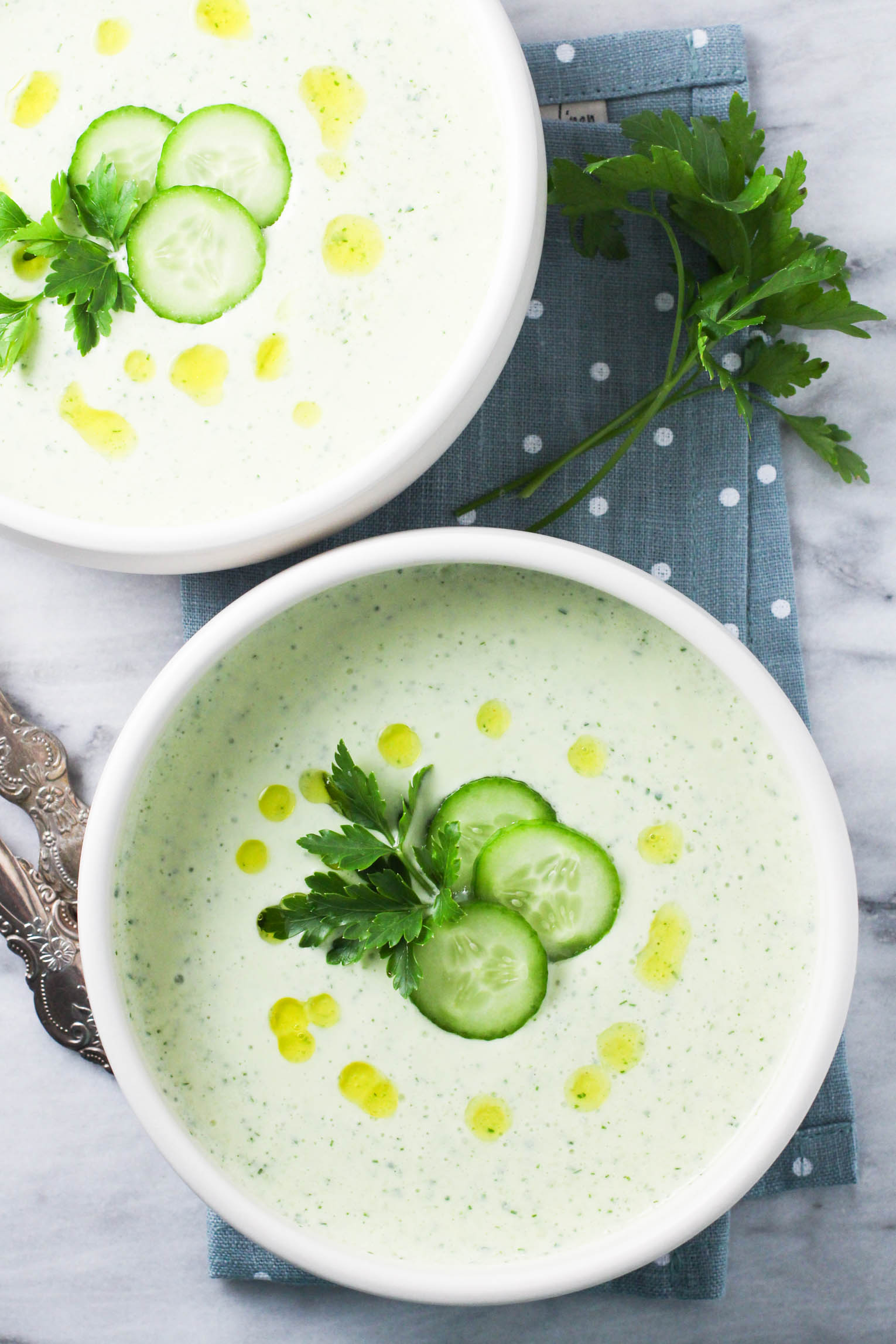 Overhead shot of two bowls of the cucumber cottage cheese soup standing on a linen napkin. The soup is garnished with cucumber slices, parsley and olive oil.