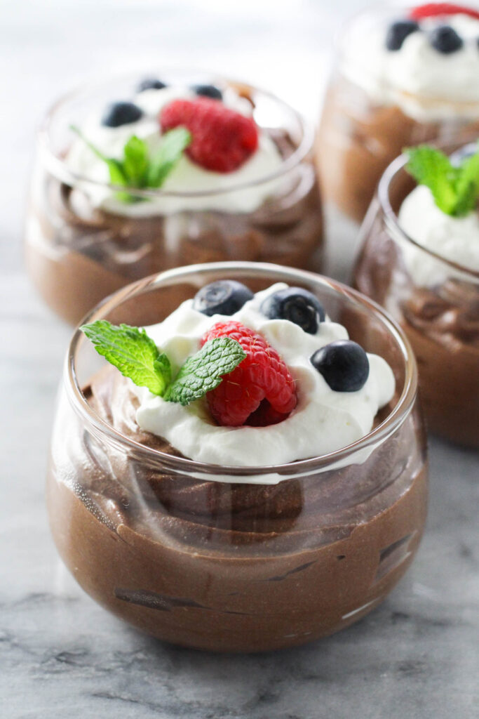 Cottage cheese chocolate mousse in glass dessert bowls garnished with whipped cream, raspberries, blueberries, and mint leaves.