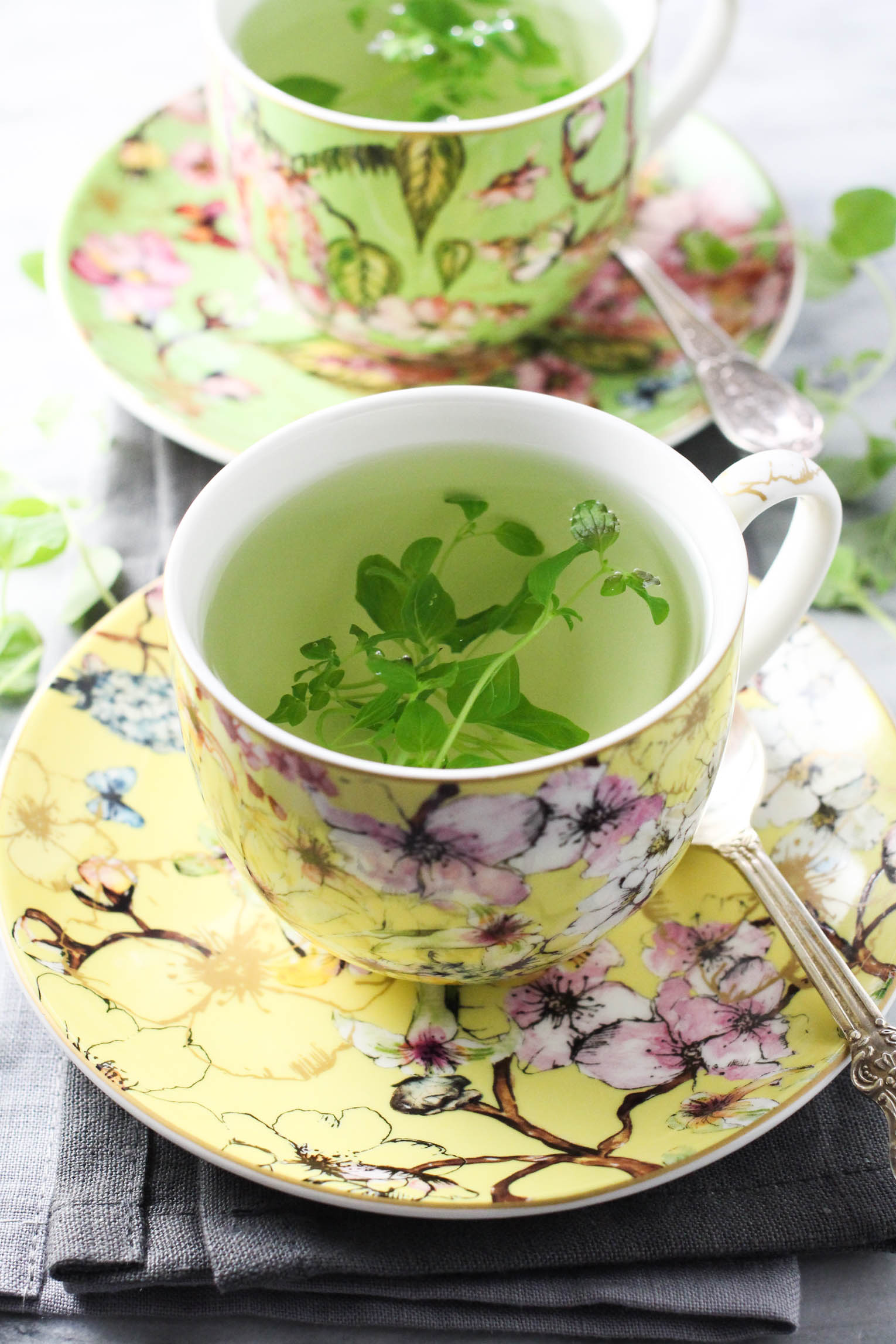 Oregano tea with fresh herbs in teacup standing on a saucer with a silver spoon to the right. Another teacup in the background.