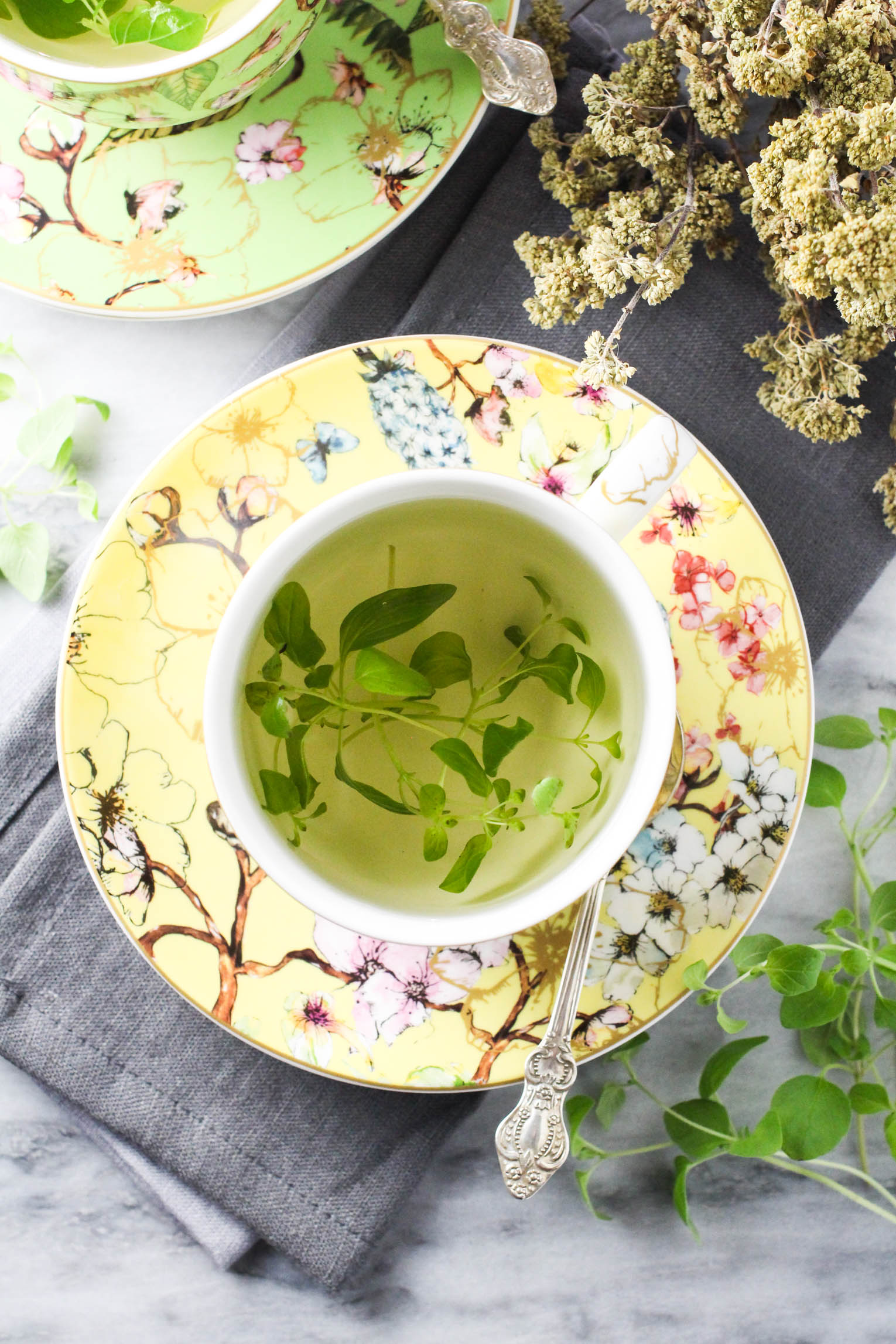Overhead shot of fresh oregano tea and a teacup standing on a saucer with a silver spoon to the right. Dried oregano in the top right corner.