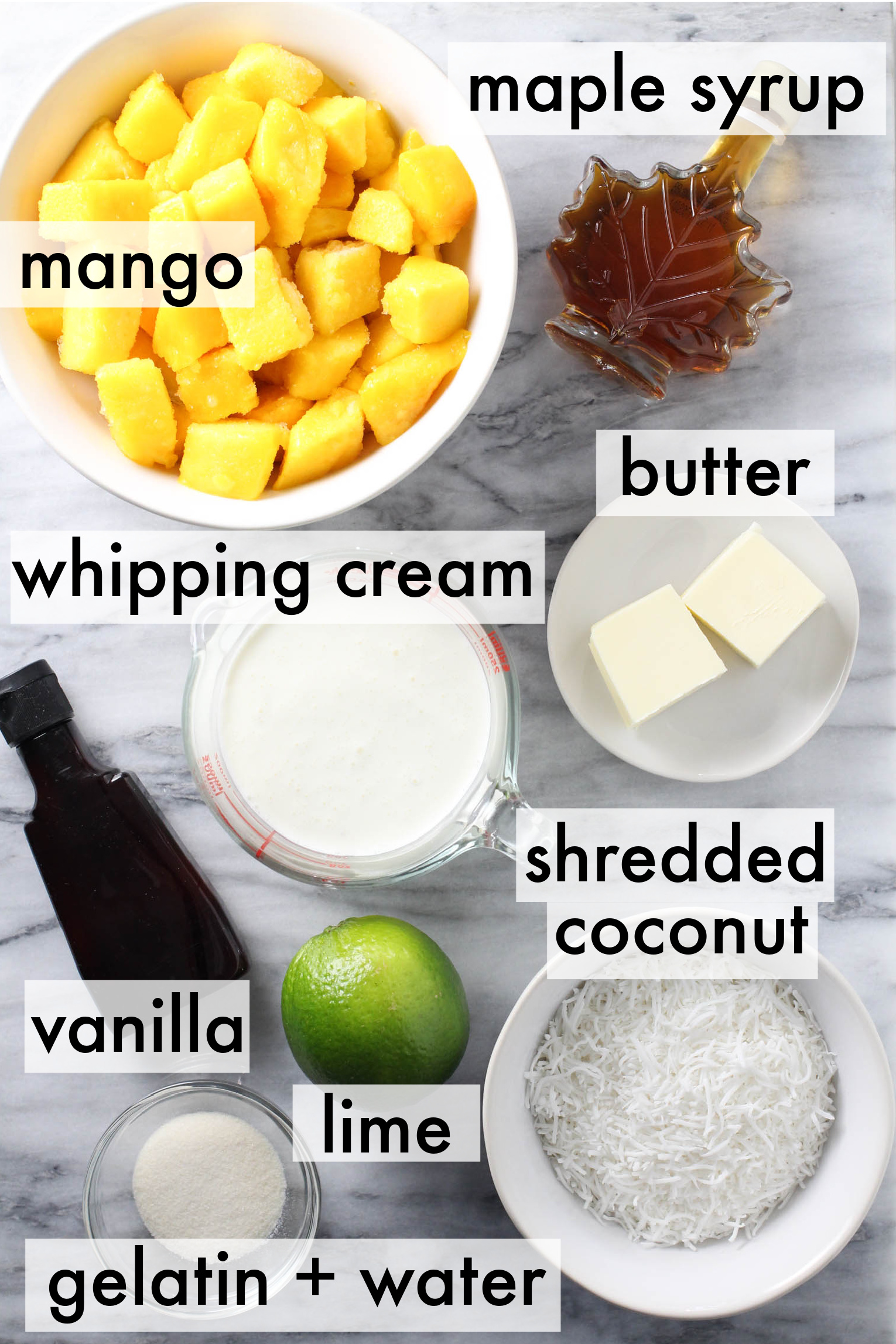 Mango mousse ingredients on marble background. The ingredients are lebeled as follows: maple syrup, mango, butter, whipping cream, shredded coconut, vanilla, lime, gelatin+water.