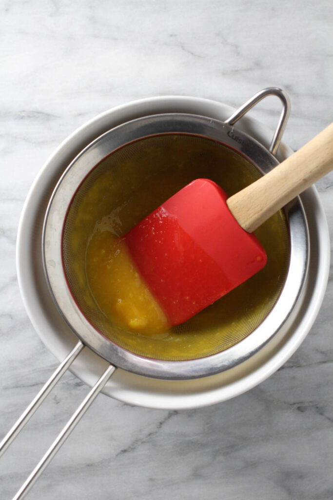 Mango puree being pushed through a fine-mesh sieve with a rubber spatula.
